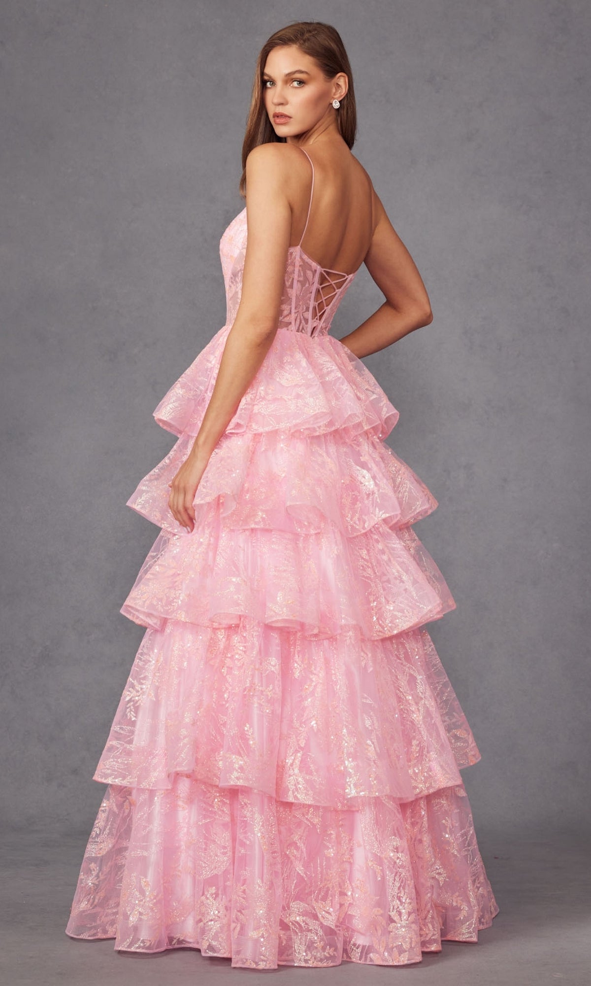 A woman wears the Long Prom Dress JT2438K by Juliet, a sleeveless pastel gown featuring thin straps, a fitted bodice, and tiered layered skirt, standing against a gray background.